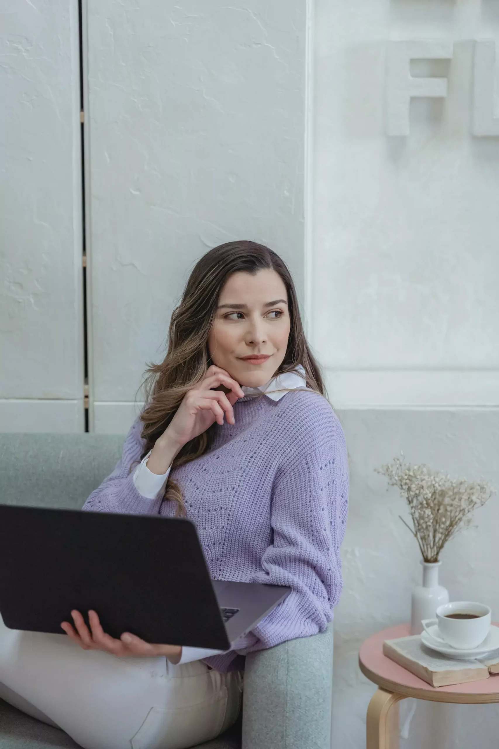 Zamyślona młoda kobieta we fioletowym swetrze trzymająca laptopa
