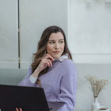 Zamyślona młoda kobieta we fioletowym swetrze trzymająca laptopa